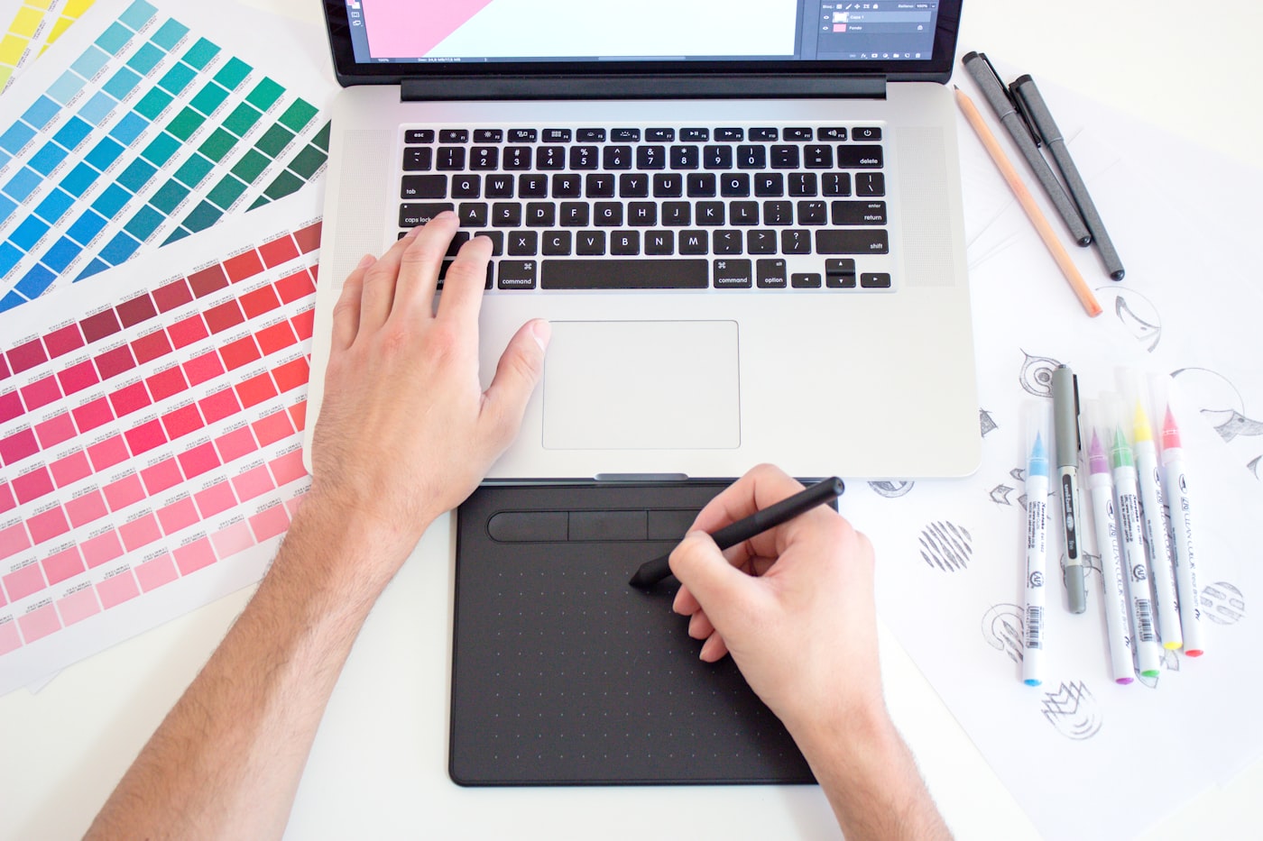 A designer's desk with colour swatches, a drawing tablet and a laptop open to a design canvas.